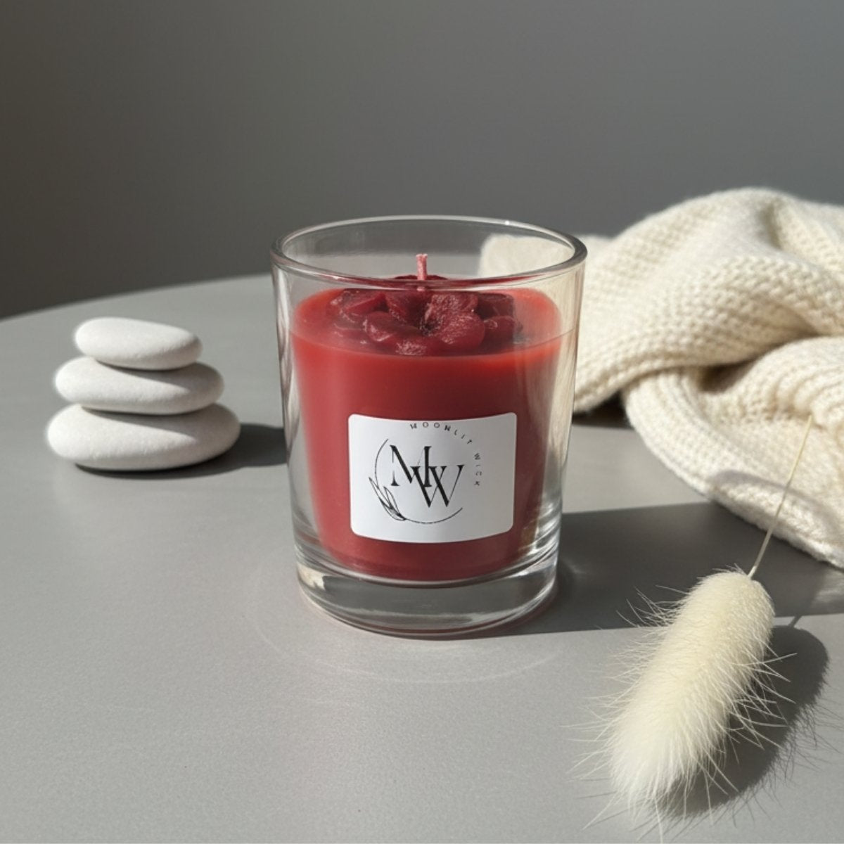 Red candle in a glass container with a label on a gray surface, surrounded by stones and a textured fabric