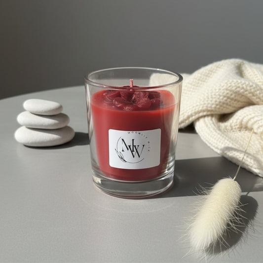 Red candle in a glass container with a label on a gray surface, surrounded by stones and a textured fabric